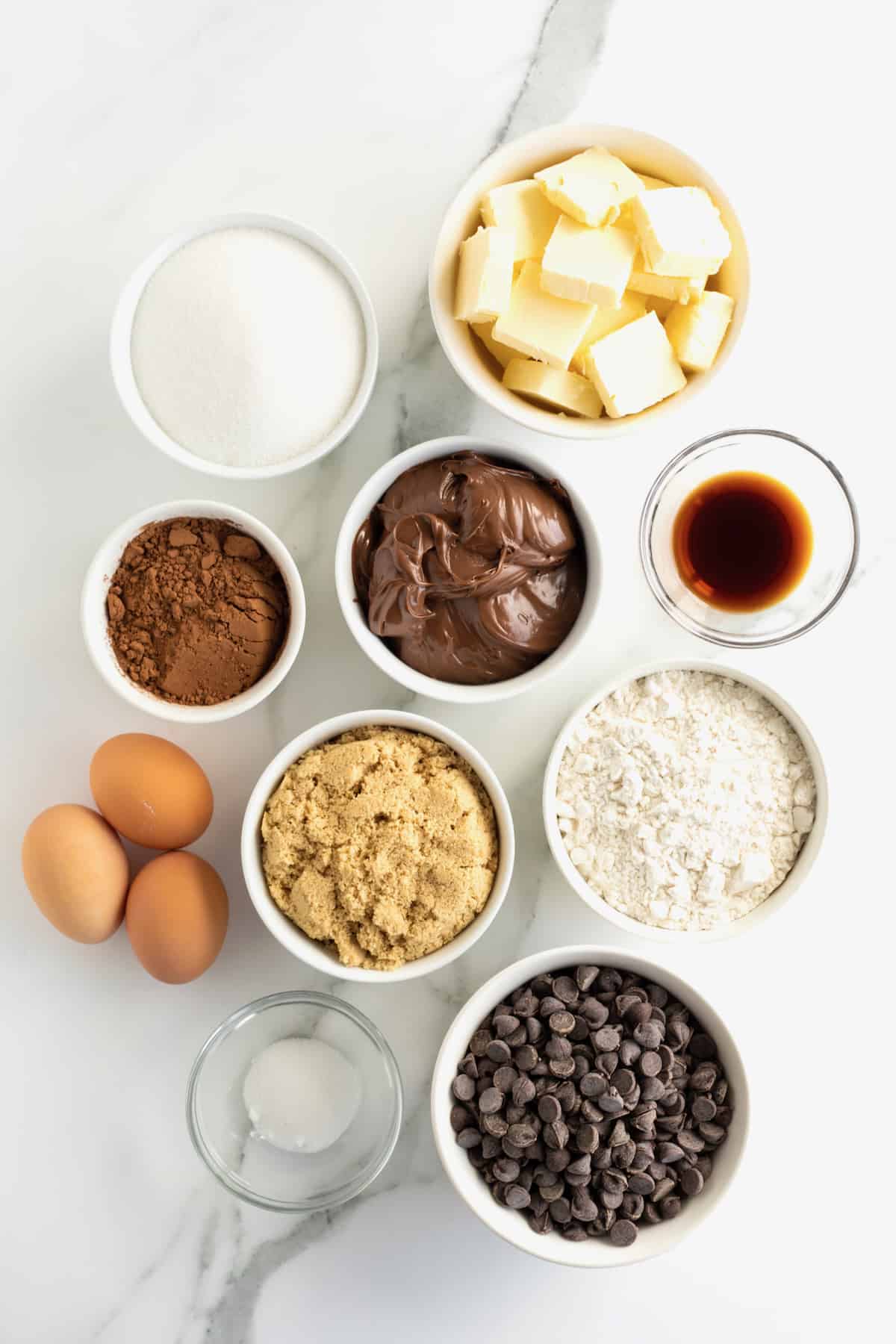 Butter, chocolate chips, granulated sugar, brown sugar, cocoa powder, vanilla, All-purpose flour, kosher salt, Nutella chocolate hazelnut spread in small containers on a white counter top next to three large brown eggs.
