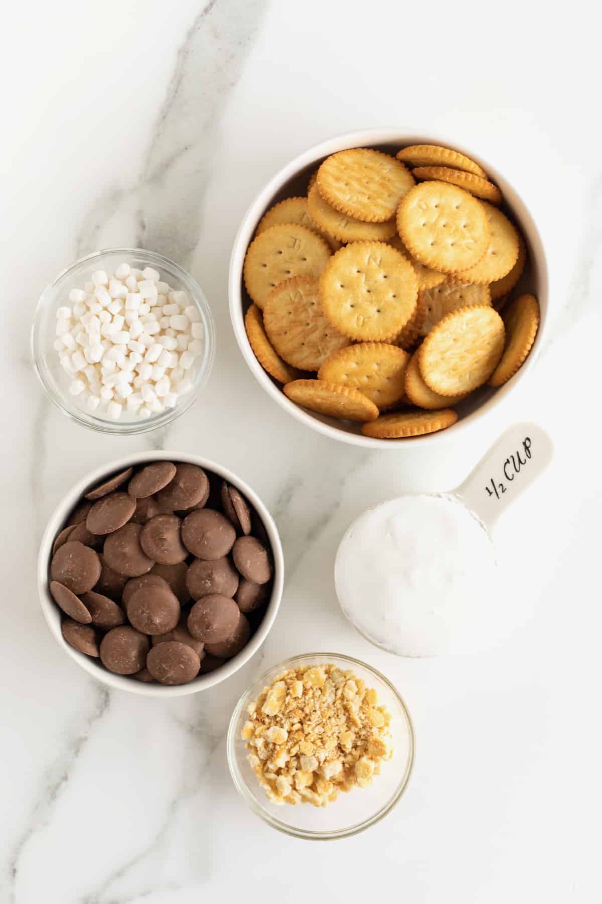 Ritz crackers, marshmallow bits, crushed cracker bits, marshmallow creme, and chocolate candy wafers in small glass dishes on a white marble counter.