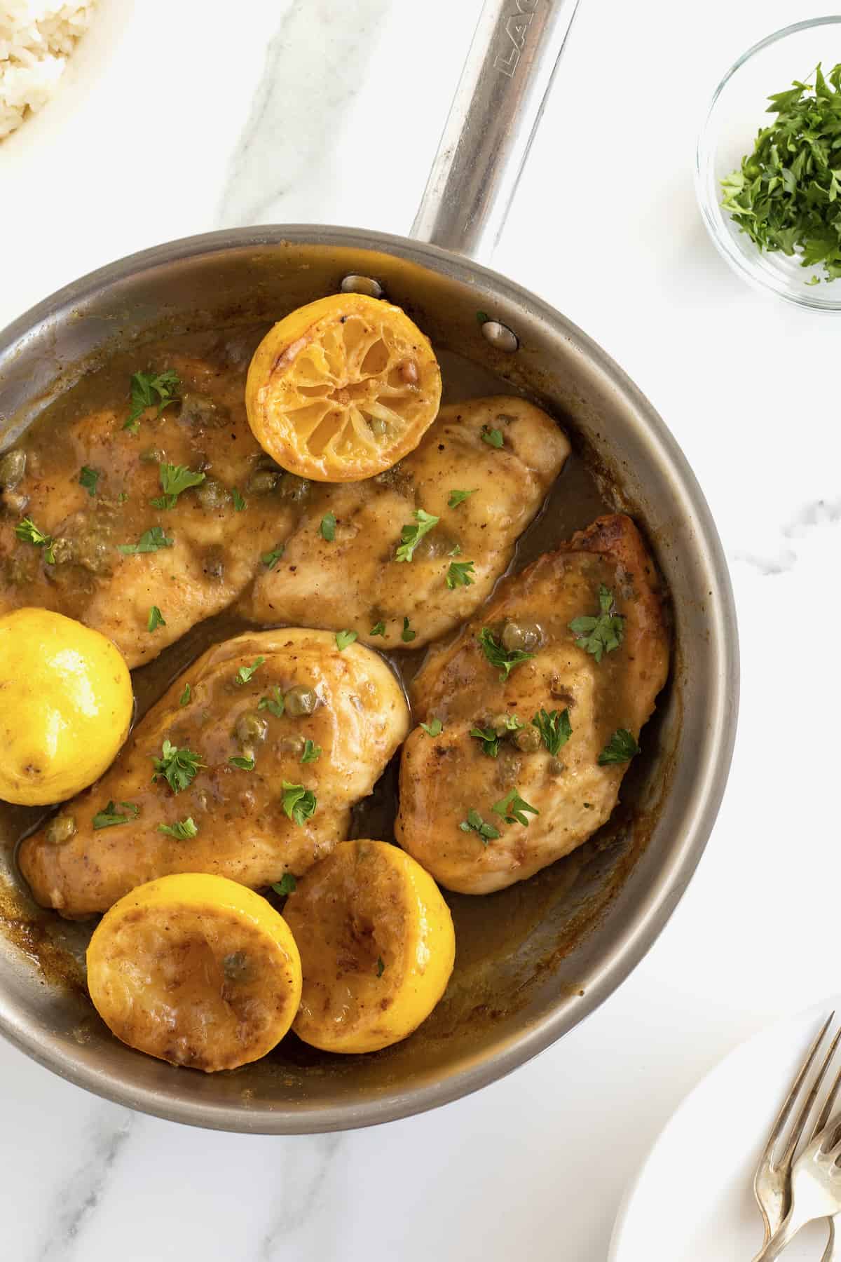A skillet with chicken breasts, halved lemons , capers and sauce.