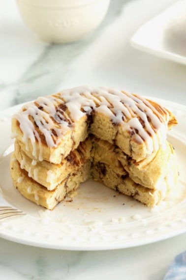 A stack of three pancakes made out of canned cinnamon rolls on a white plate drizzled with icing.