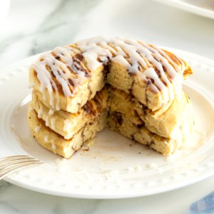 A stack of three pancakes made out of canned cinnamon rolls drizzled with icing on a white plate.