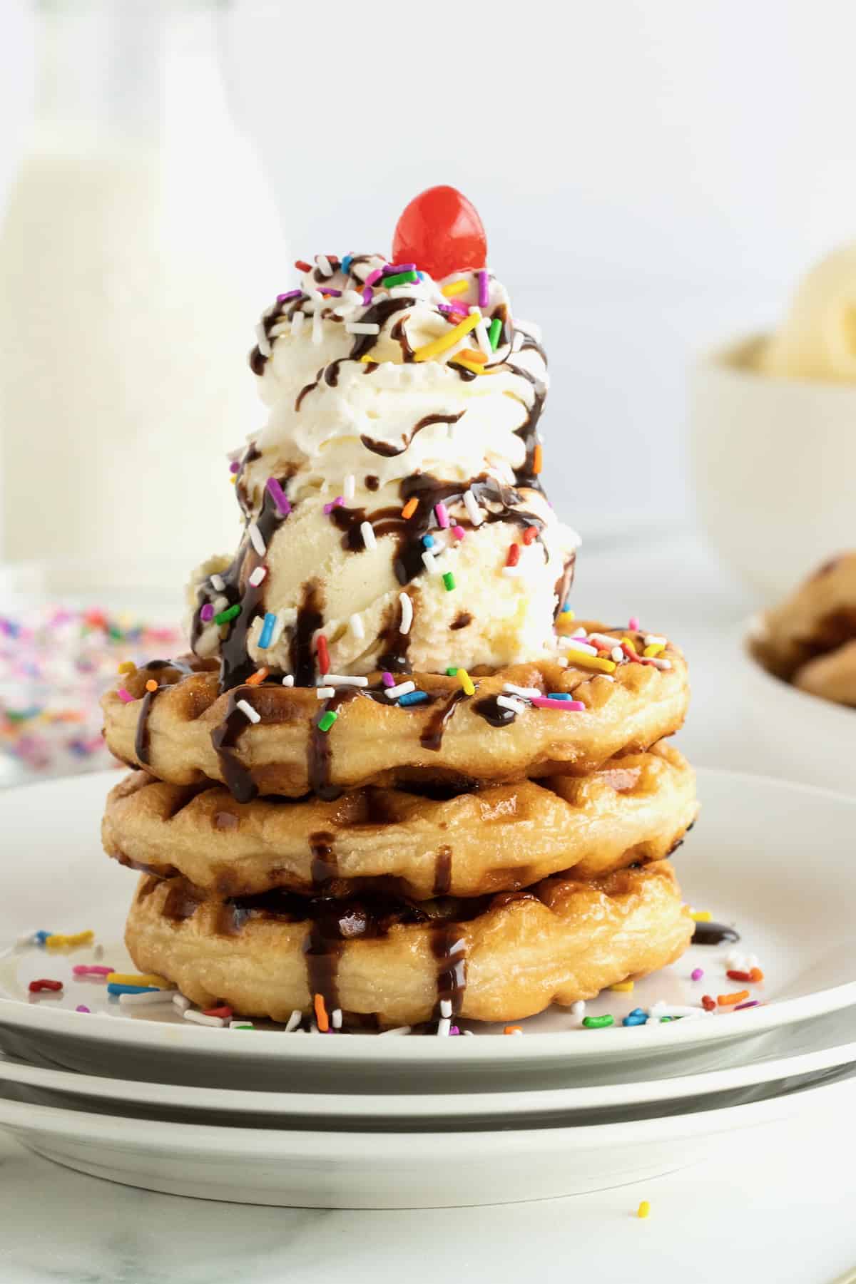 An ice cream sundae made with a stack of three donut waffles, two scoops of vanilla ice cream, chocolate sauce, rainbow sprinkles and a cherry on top.