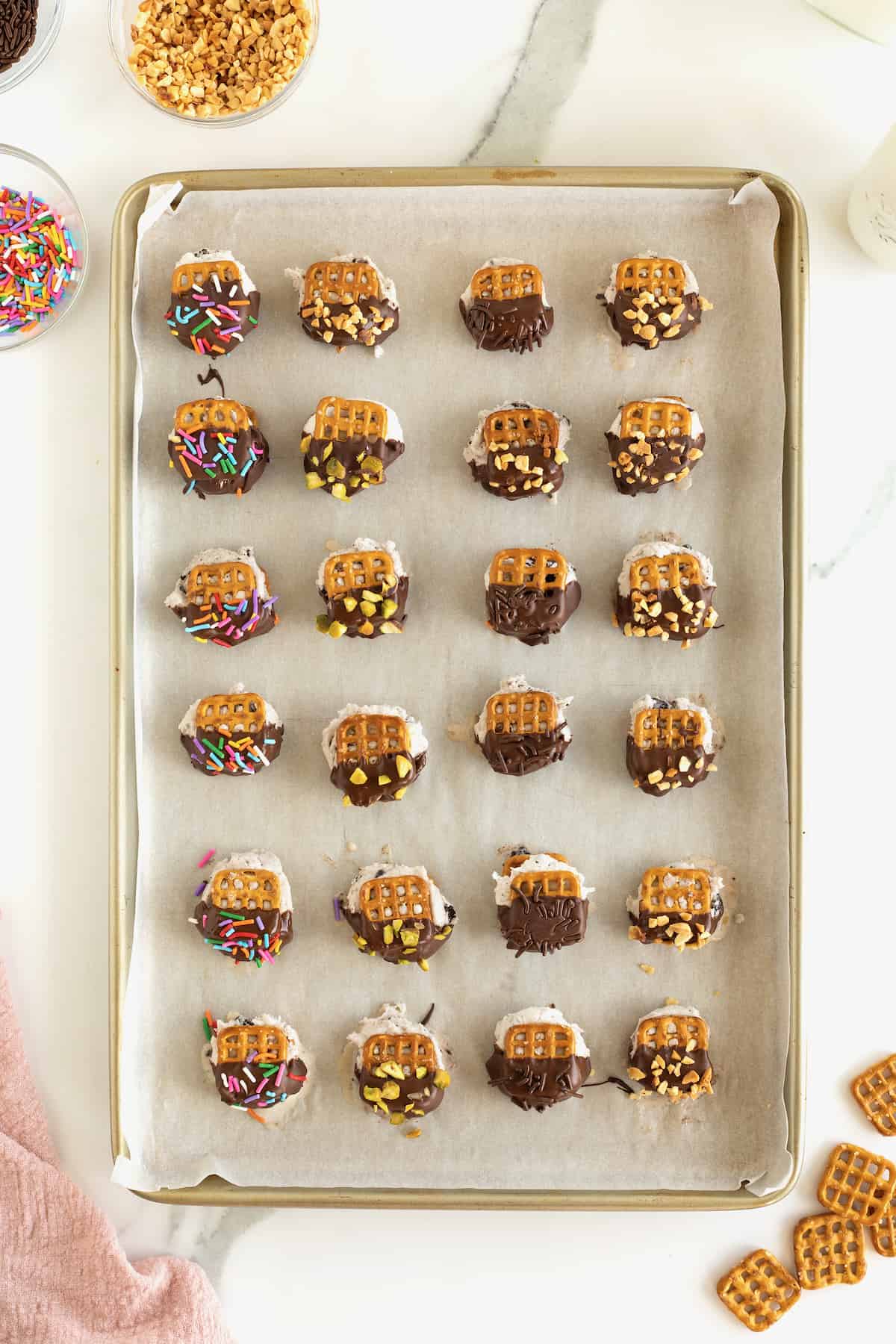A parchment lined baking tray filled with pretzel snap ice cream bites dipped in chocolate and coated in sprinkles and chopped nuts.