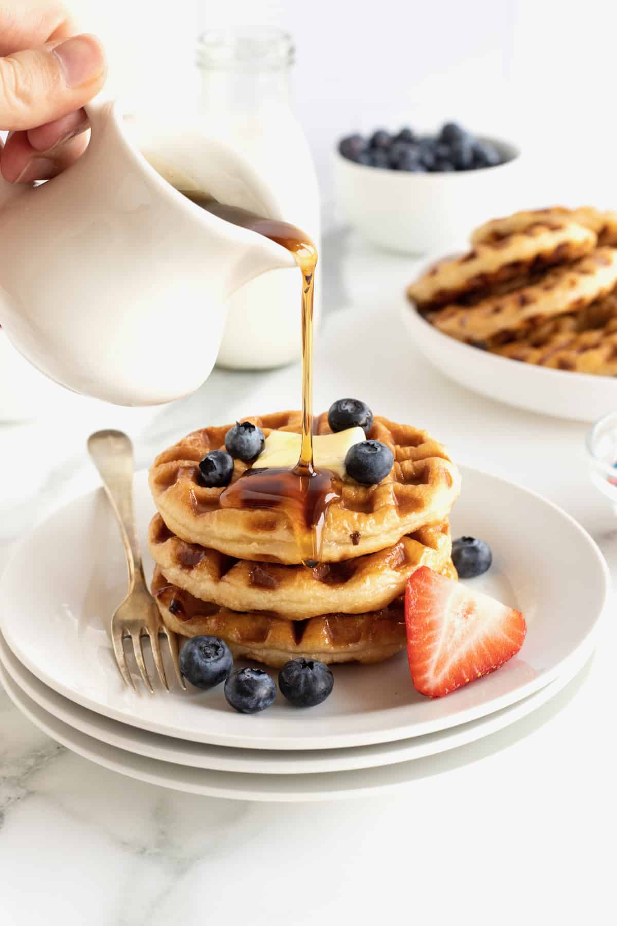 Three donut waffles stacked on a white plate with a square of butter, fresh blueberries and hot maple syrup poured over the top.