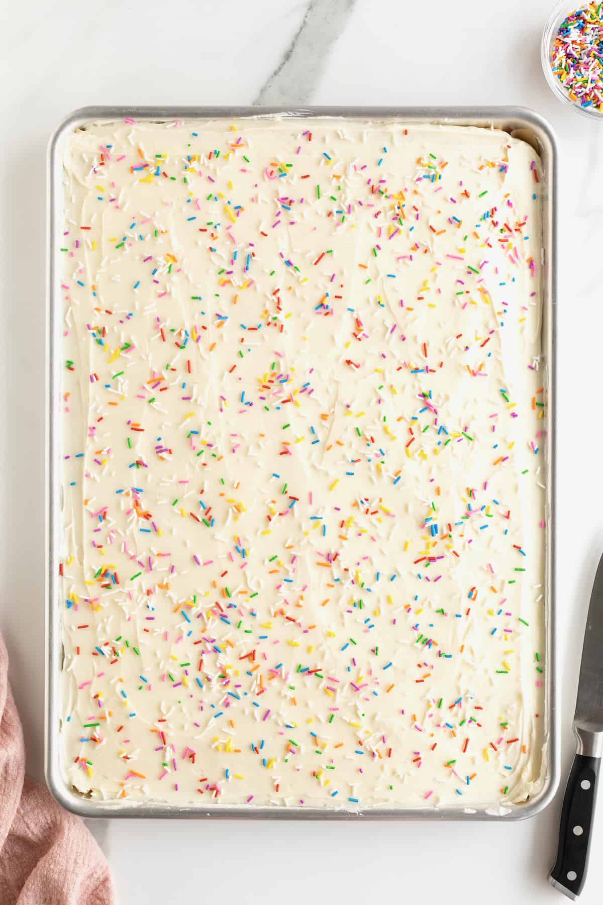 A sheet pan of cookies covered with white frosting and rainbow sprinkles.