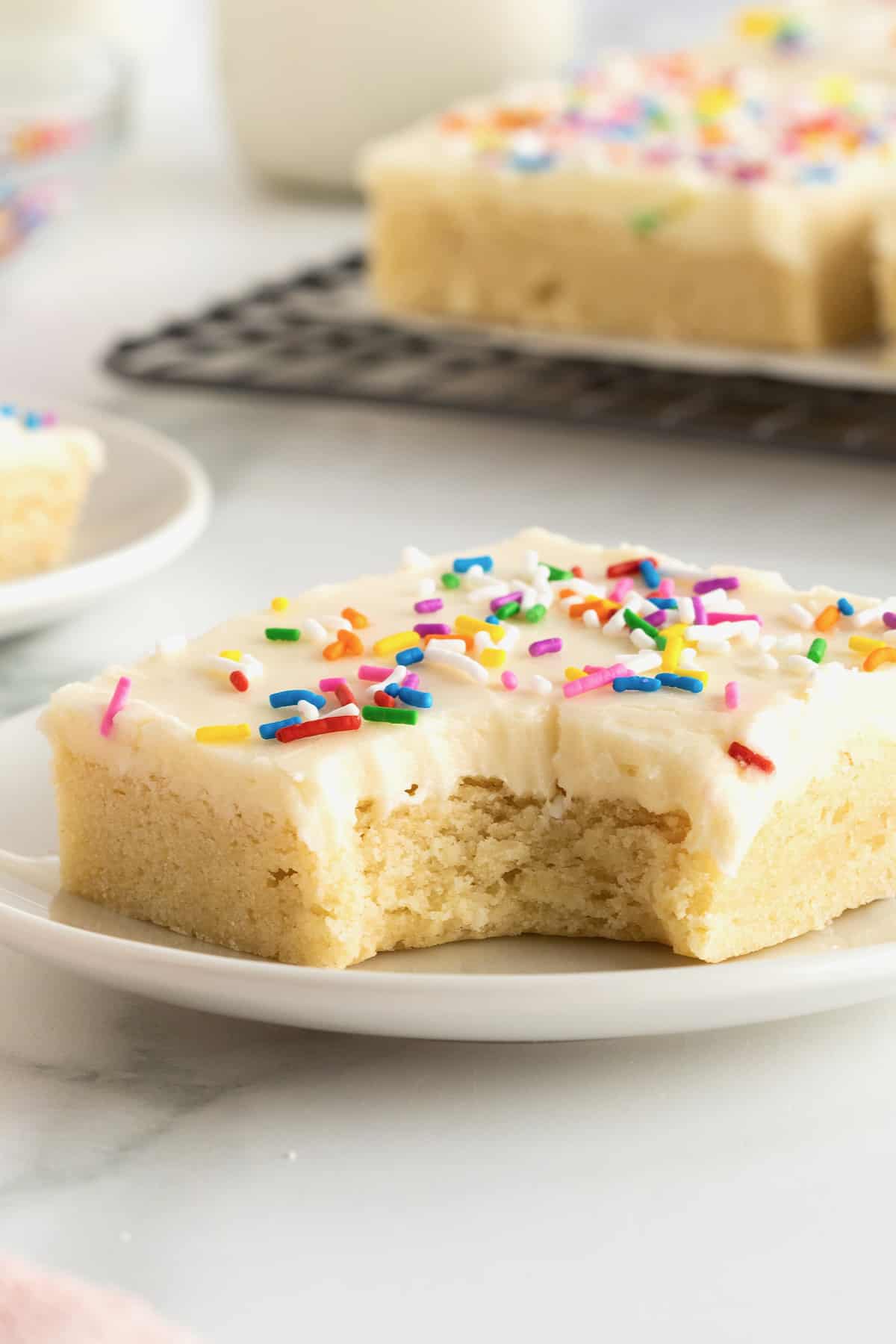 A square sugar cookie with frosting and rainbow sprinkles on a white plate.