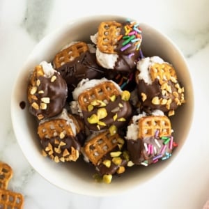 A white serving bowl filled with pretzel ice cream sandwich bites dipped in chocolate and coated in sprinkles.
