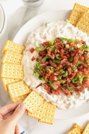 A white serving bowl with a creamy white dip topped with crumbled bacon and shredded lettuce with club crackers on the side.
