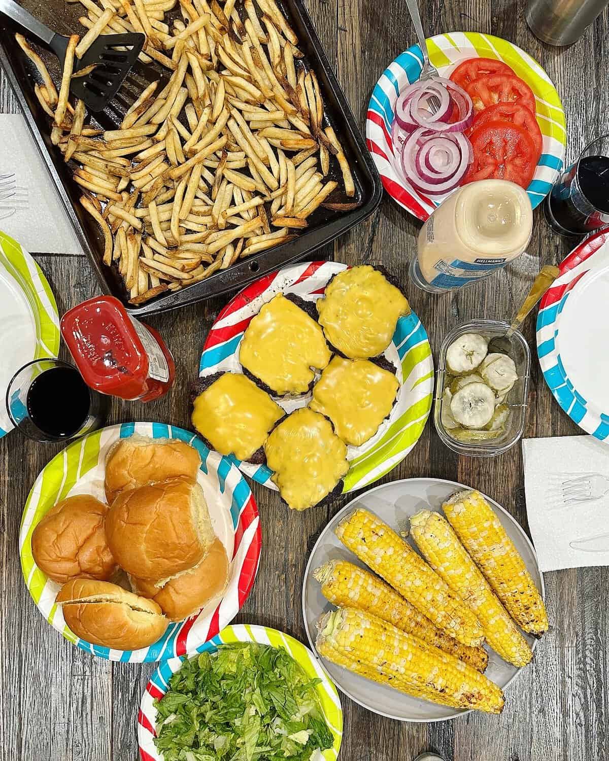 A baking sheet of French fries, a plate of five cheese burgers with yellow cheese, a plate of buns and a plate of grilled corn on a wooden table.
