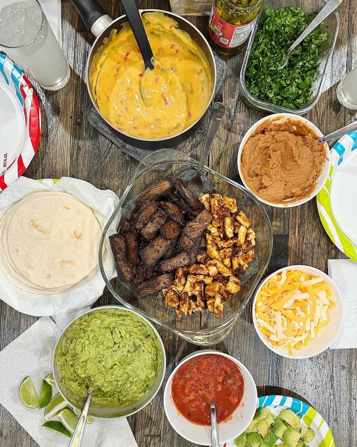 Fajita chicken and steak in a glass bowl surrounded by tortillas and bowls of queso, refried beans, guacamole, shredded cheese and salsa.