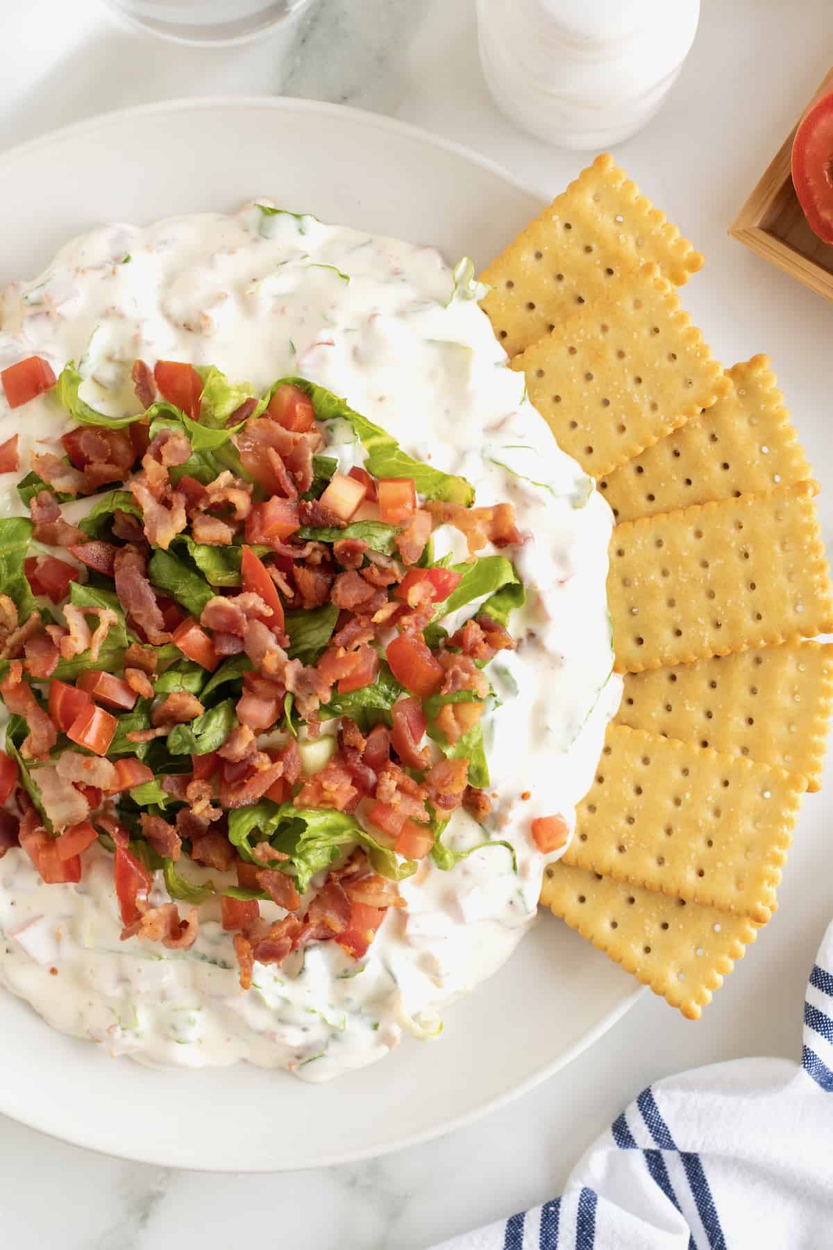 A white serving bowl with a creamy white dip topped with crumbled bacon and shredded lettuce with club crackers on the side.