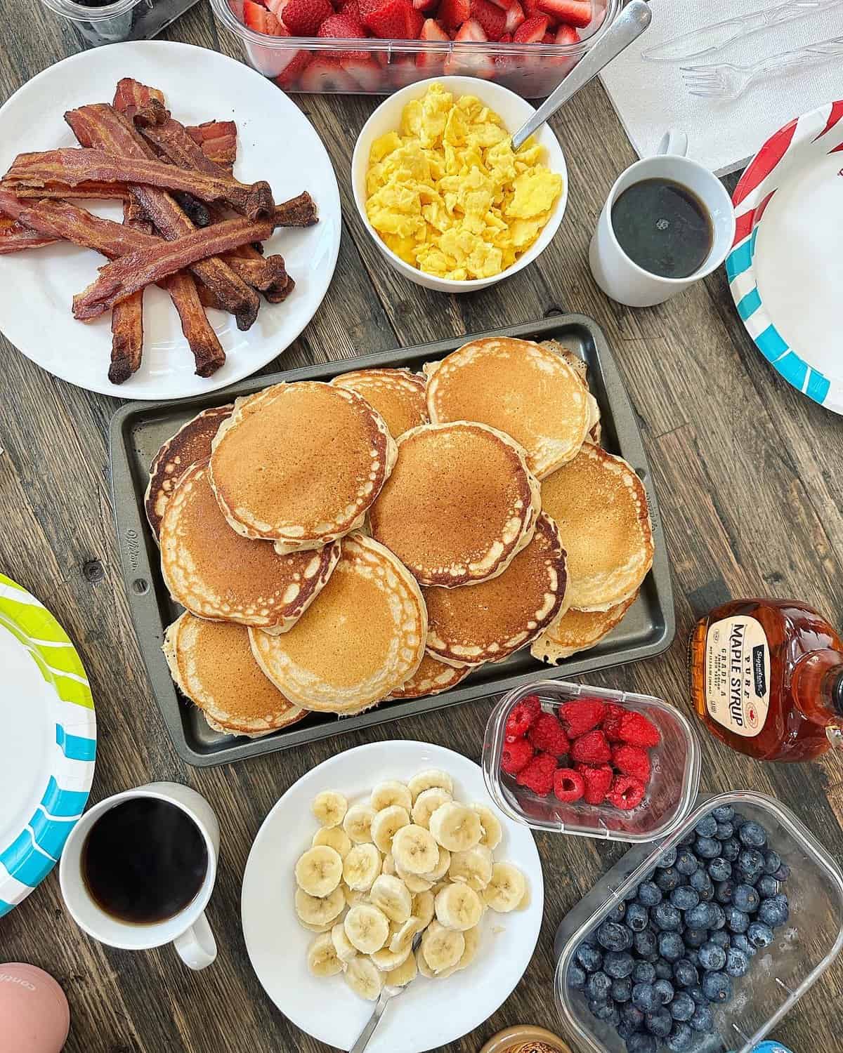 Baked bacon on a white plate, scrambled eggs in a white serving bowl, a baking sheet filled with pancakes and two cups of coffee on a wooden table.