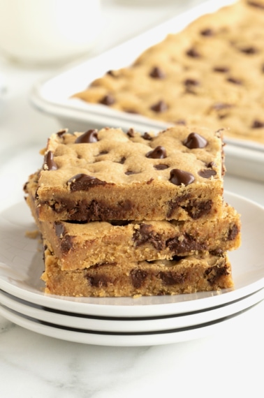 A stack of three peanut butter cookie bars with chocolate chips on a stack of three white plates with a silver sheet pan of cookie bars partially visible in the background.