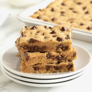 A stack of three peanut butter cookie bars with chocolate chips on a stack of three white plates with a silver sheet pan of cookie bars partially visible in the background.