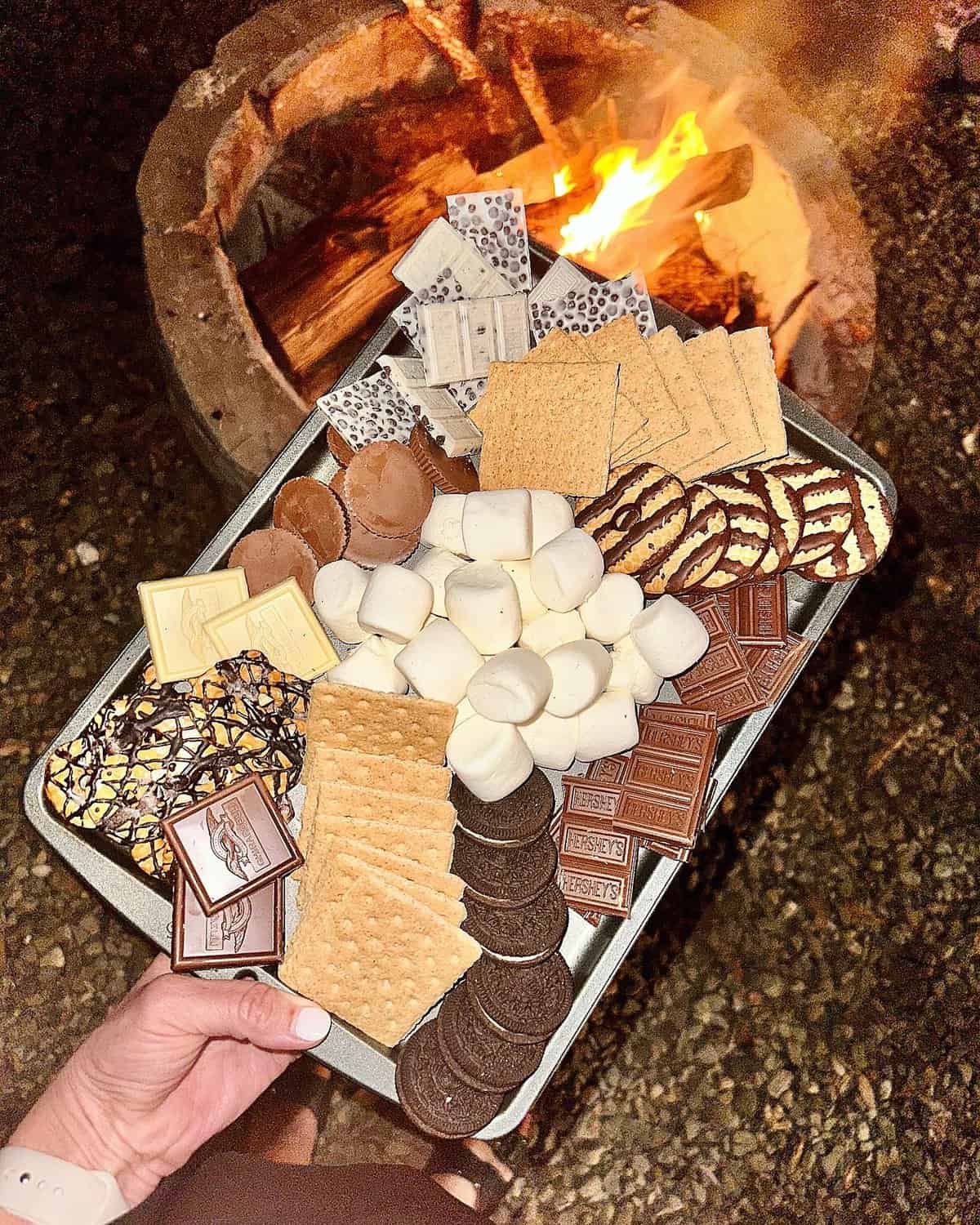 A baking tray filled with marshmallows, graham crackers, chocolate, fudge striped cookies in front of a fire pit.