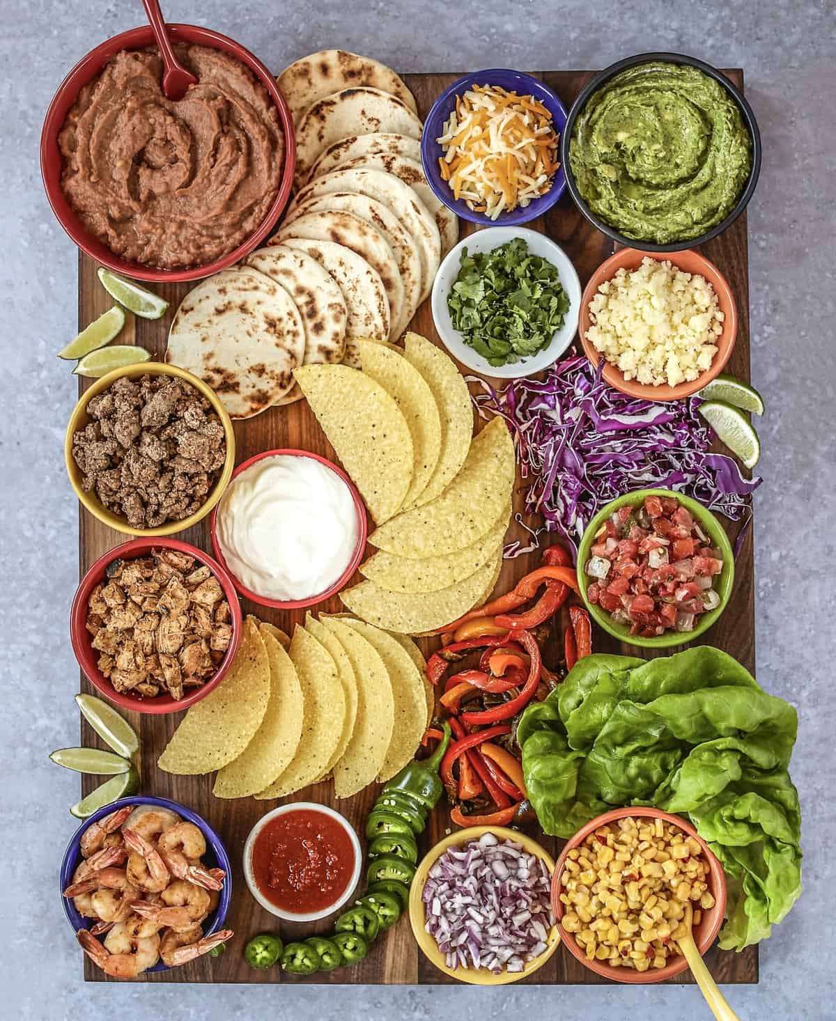 A large rectangle wood board with taco shells, flour tortillas, meats, shredded cheese, guacamole and refried beans in small containers.