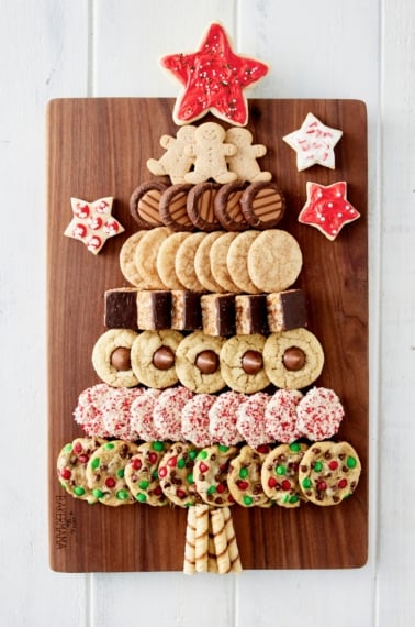 A variety of holiday cookies laid out on a rectangular wood food board in the shape of a Christmas tree with a red star cookie on top.