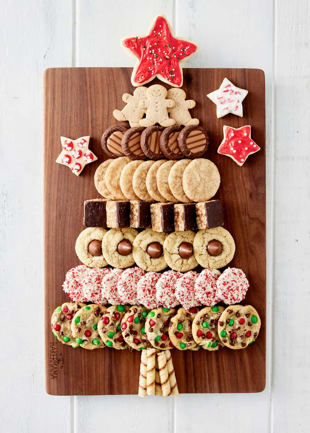 A variety of holiday cookies laid out on a rectangular wood food board in the shape of a Christmas tree with a red star cookie on top.