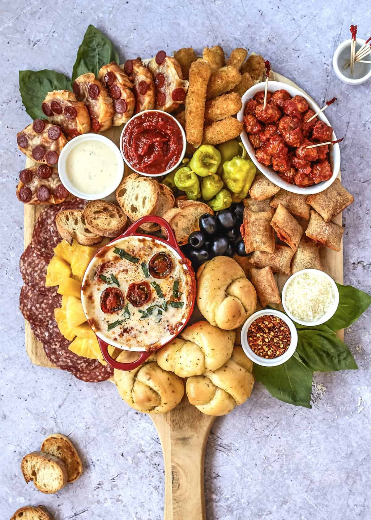 A large round wood board with garlic knots, pizza pockets, pizza bagels, mozarella sticks, toasted baguette slices, and peppers along with olives.