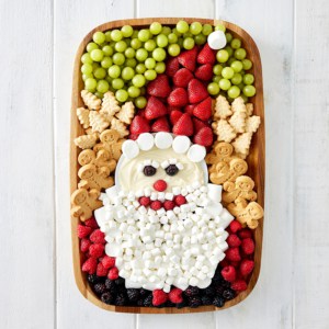 A wood food board decorated with fruit and cookies and marshmallows to look like Santa.