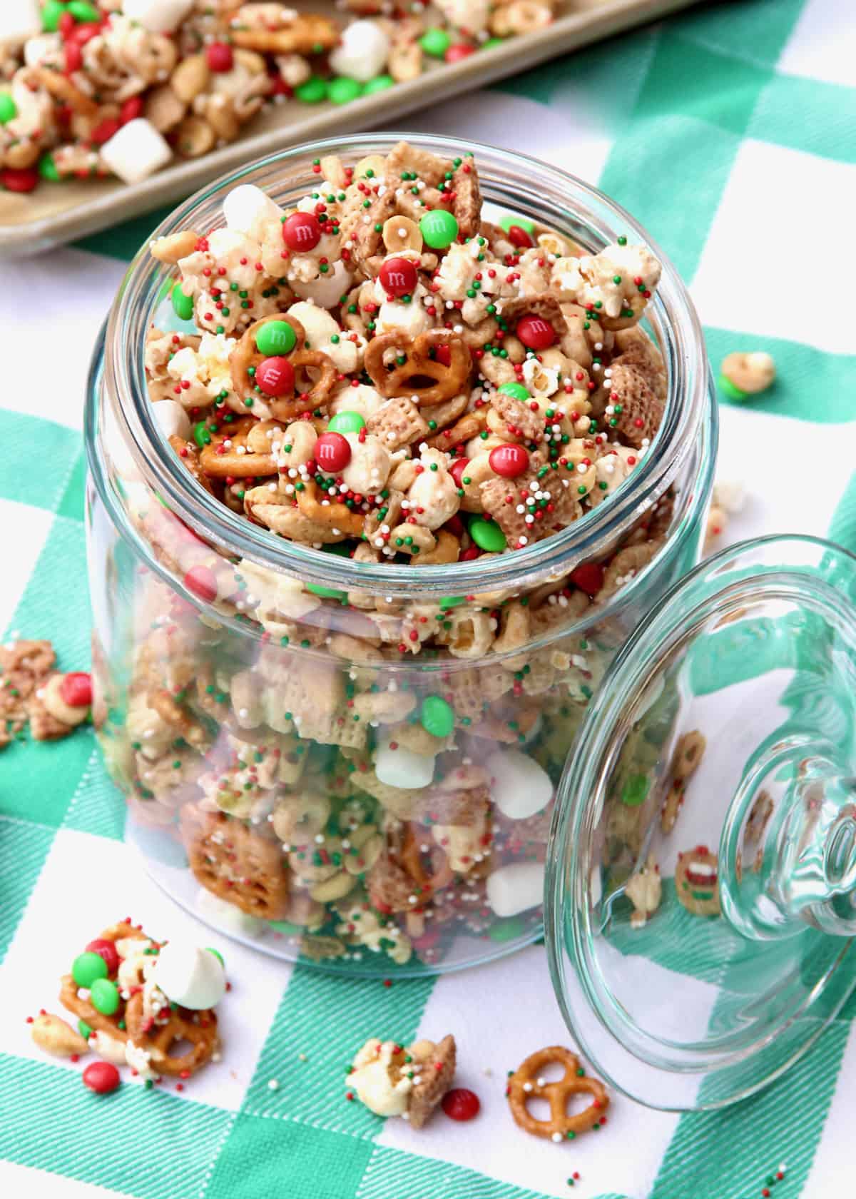 A jar of holiday party snack mix on a green and white gingham table cloth.
