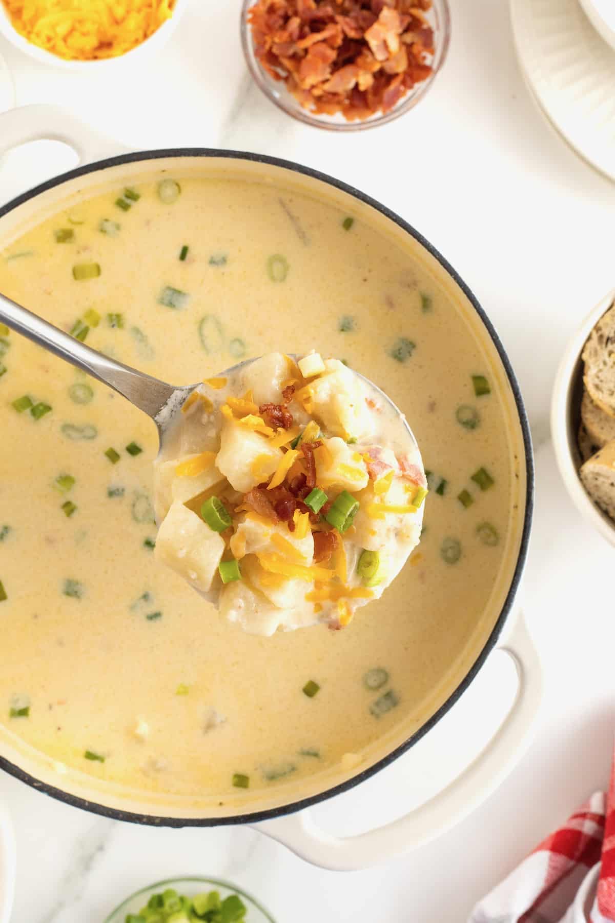 A silver ladle full of potato soup over a white ceramic coated pot of baked potato soup.
