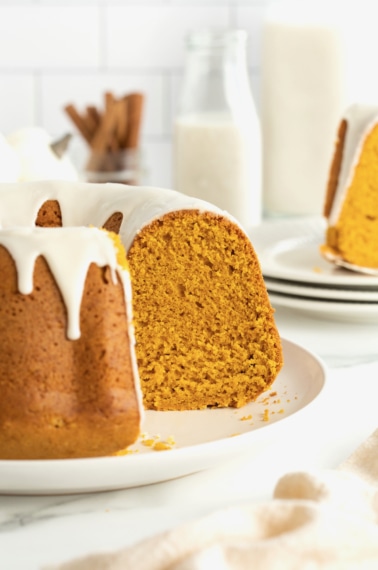 A pumpkin pound cake topped with a white glaze dripping down it on a white serving platter. One slice is missing from the cake.