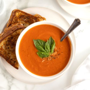 A bowl of homemade tomato soup next to two triangle cut halves of a grilled cheese sandwich on a large white round plate.
