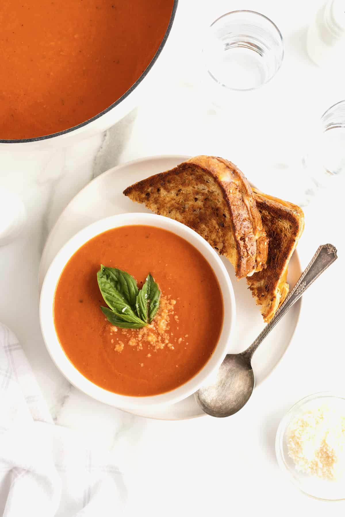 A white bowl of tomato soup on a large white plate with two triangle grilled cheese sandwiches.