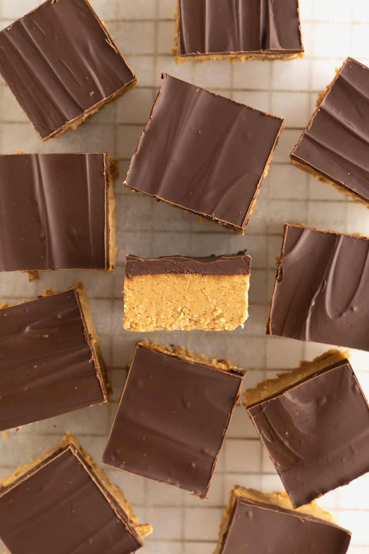 Chocolate covered peanut butter bars cut into squares  scattered on a parchment lined cooling rack. One square is turned on its side revealing the two layers.