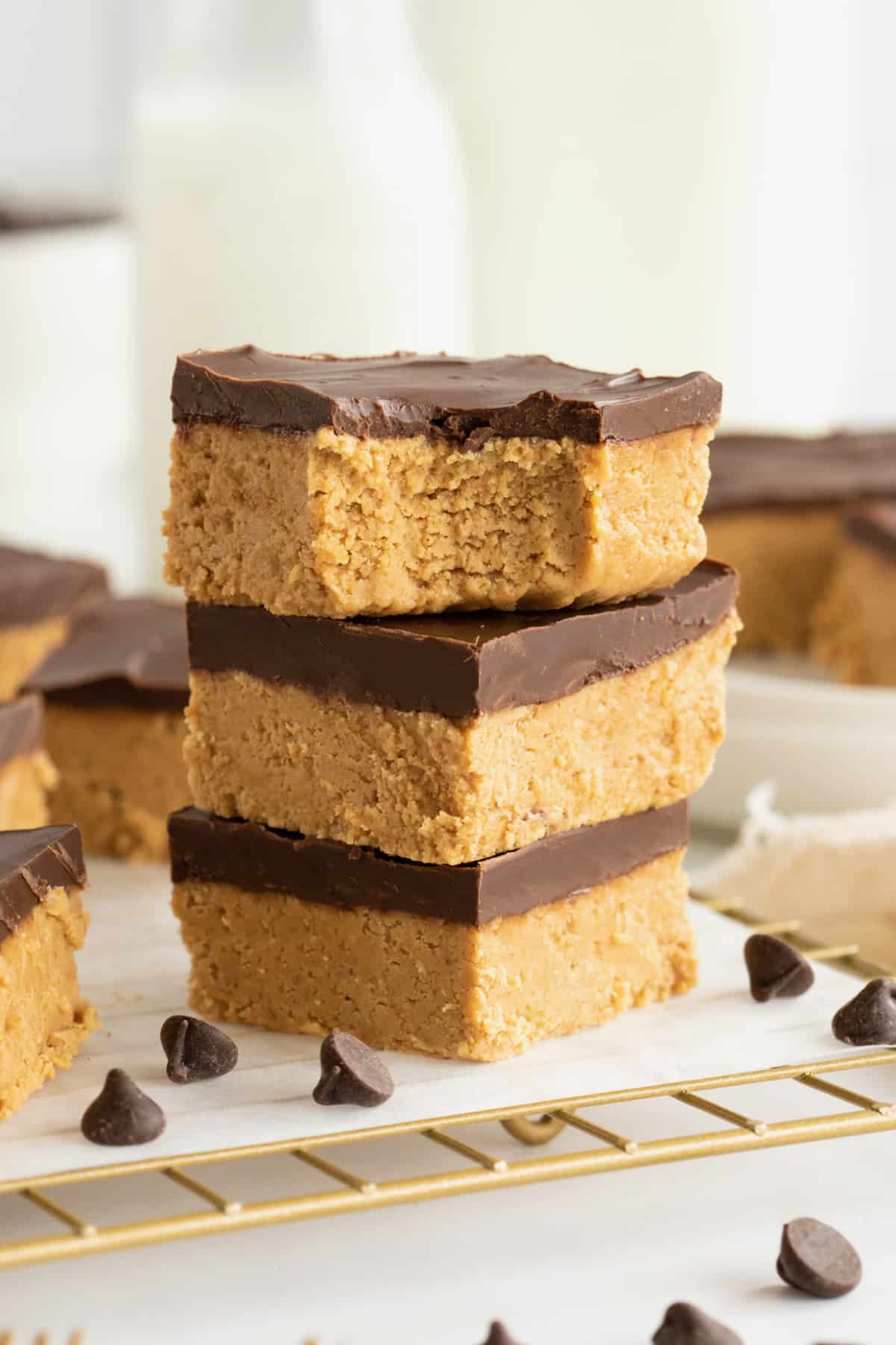 Three chocolate peanut butter bars stacked on a parchment lined brass cooling rack with chocolate chips scattered around them. The top bar has a bite out of it. 