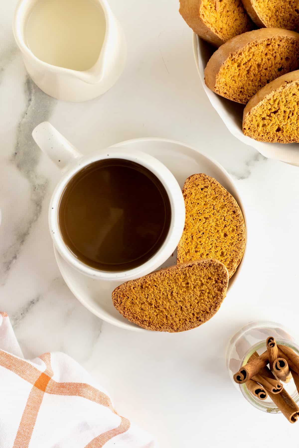 A white cup of coffee on a white saucer with two pieces of pumpkin biscotti.