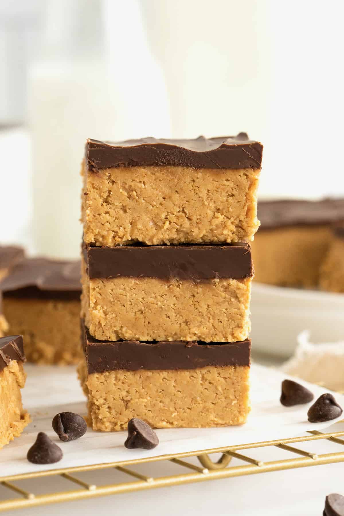 Three chocolate peanut butter bars stacked on a parchment-lined gold tone cooling rack with chocolate chips scattered around them.