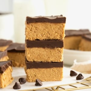 Three chocolate peanut butter bars stacked on a parchment lined brass cooling rack with chocolate chips scattered around them.