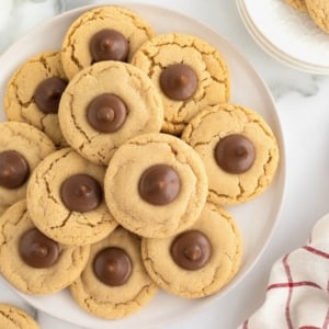 A pile of peanut butter holiday cookies with a Hershey's kiss in the center on a round white serving plate.