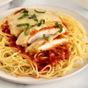 Chicken parmesan over marinara sauce and a bed of angel hair pasta on a white dinner plate.