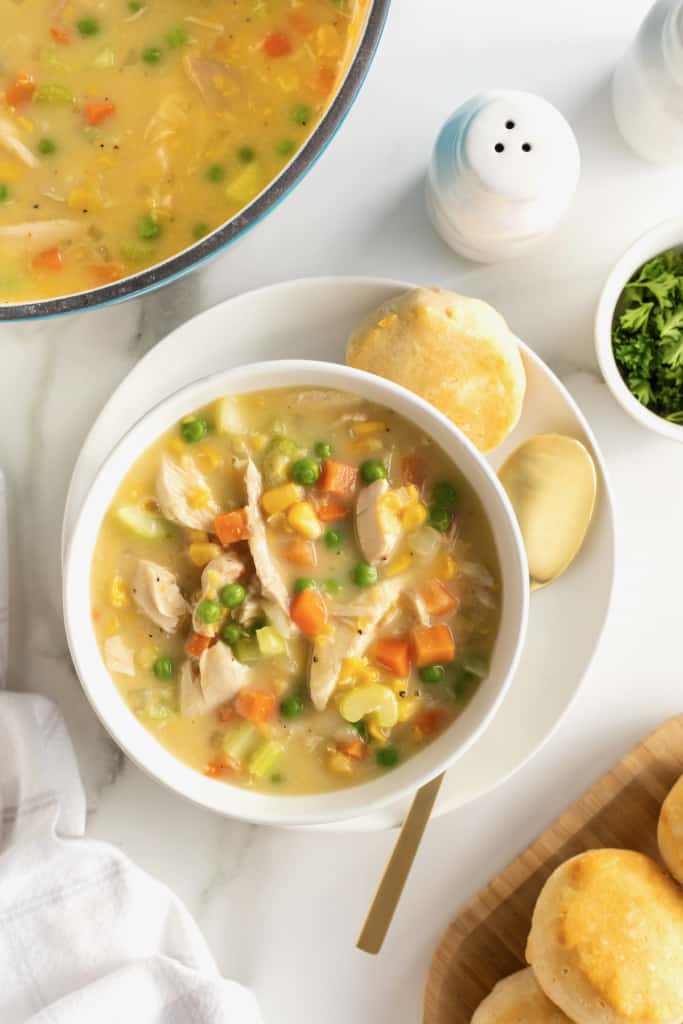 A while ceramic bowl of chicken pot pie soup on a white plate with a spoon.