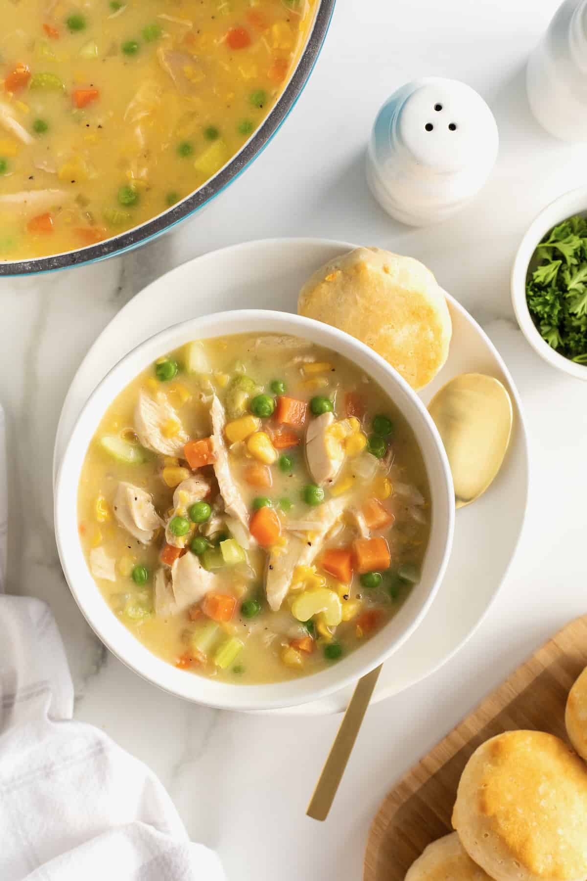 A while ceramic bowl of chicken pot pie soup on a white plate with a spoon. A biscuit is next to the bowl of chicken pot pie soup. There is a salt shaker to the upper right corner.
