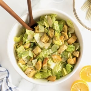 A large white serving bowl filled with caesar salad topped with croutons. Two wooden spoons stick out from under the salad.