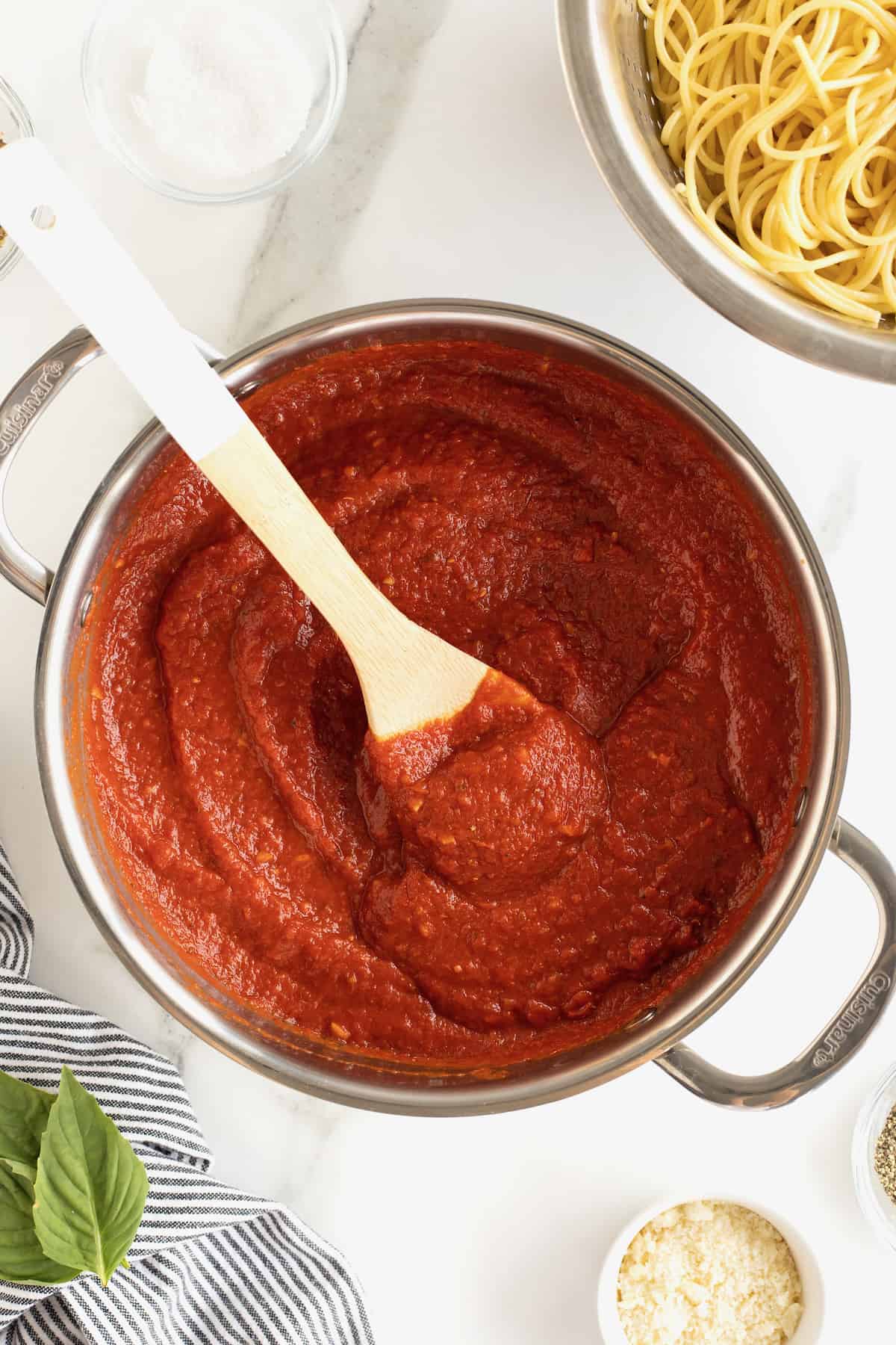 A large pot with red marinara sauce in it. A wooden spoon with a white handle is sticking out of the pot.