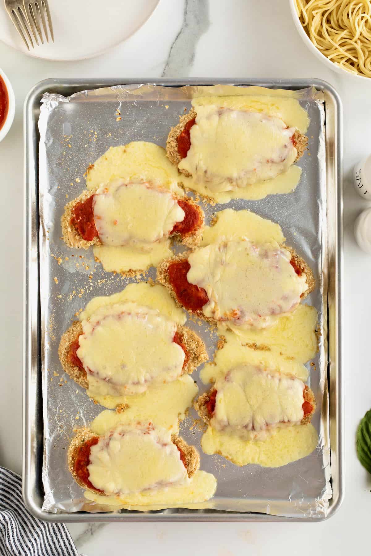 Six chicken cutlets topped with marinara sauce and melted fresh mozzarella on an aluminum baking sheet.