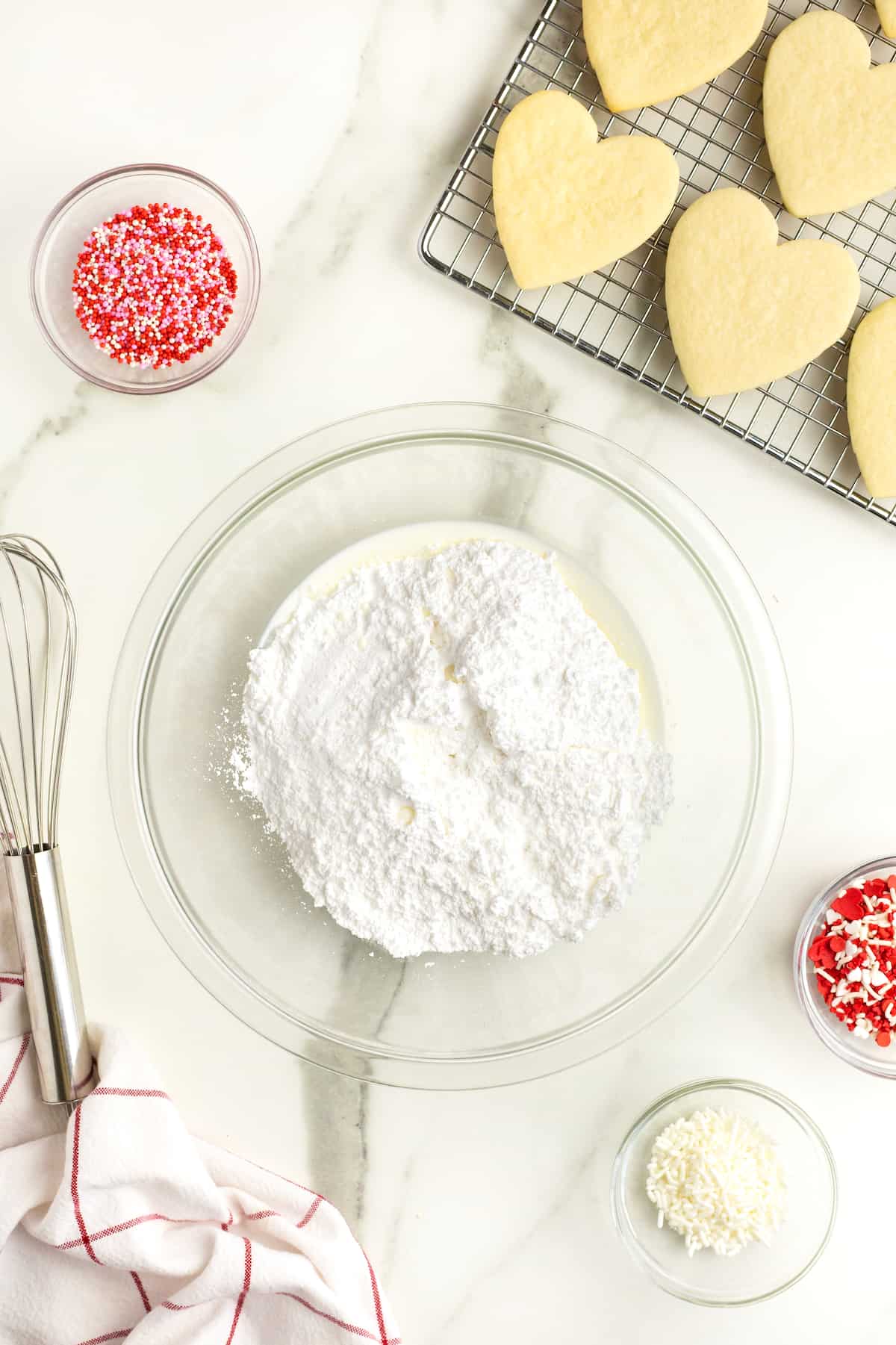 A glass bowl of powdered sugar and milk next to a wire cooling rack with heart shaped sugar cookies. A wire whisk is next to the bowl on the white marble counter.