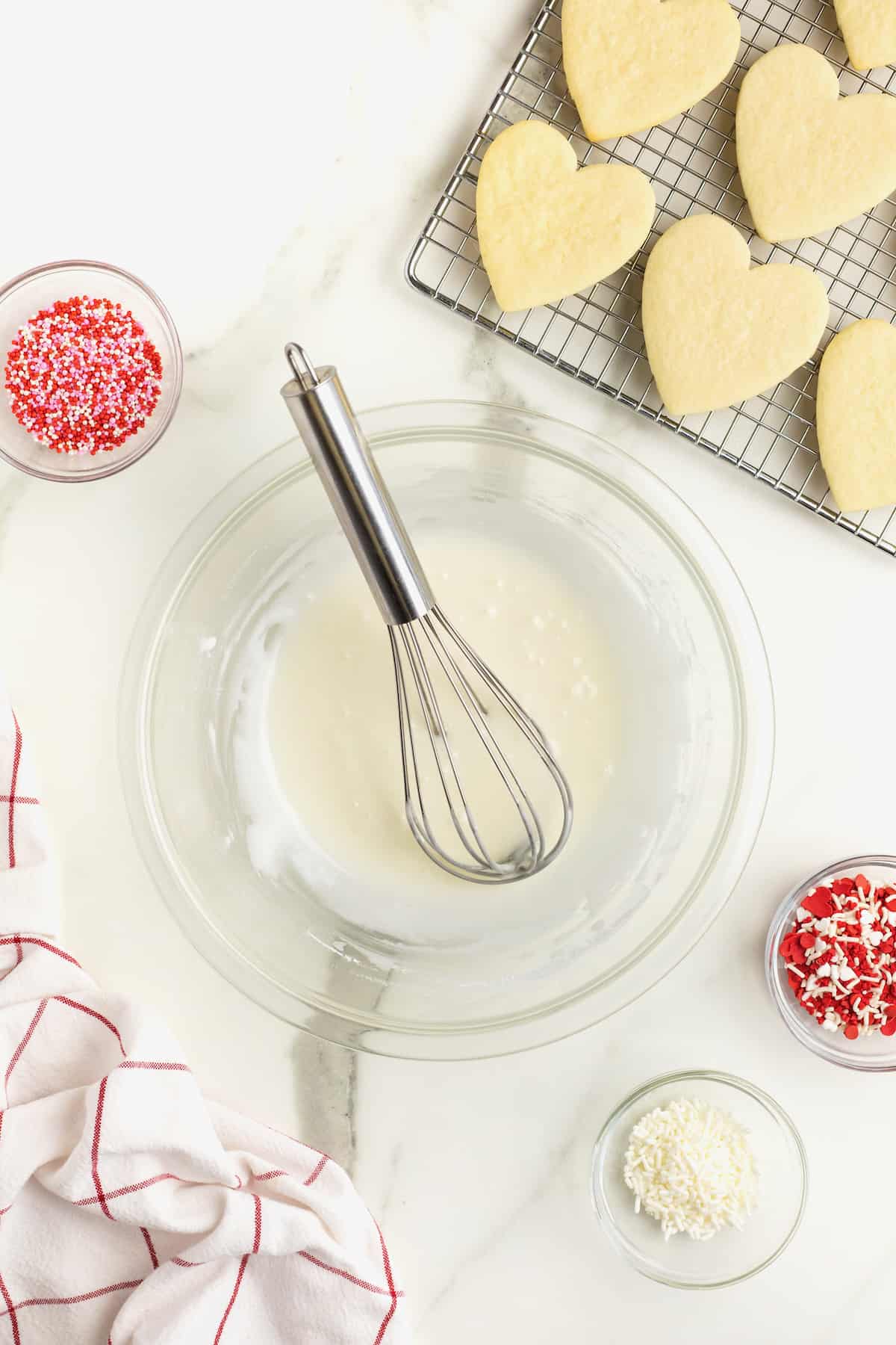 A glass bowl of icing next to a wire cooling rack with heart shaped sugar cookies on the white marble counter. There's a wire whisk in the bowl.