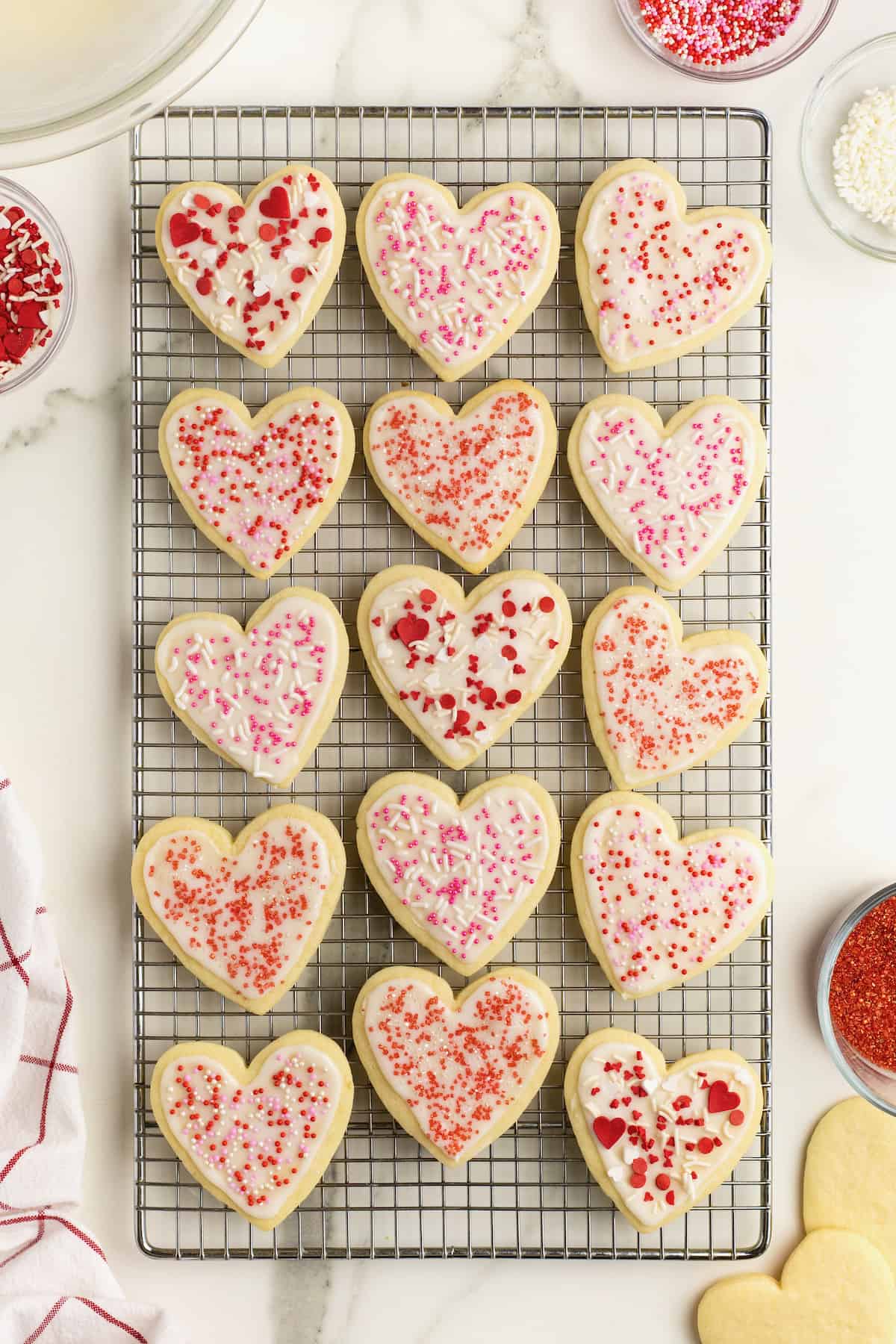 15 heart shaped sugar cookies with white frosting and red and pink sprinkles on a wire cooling rack on a white marble counter.
