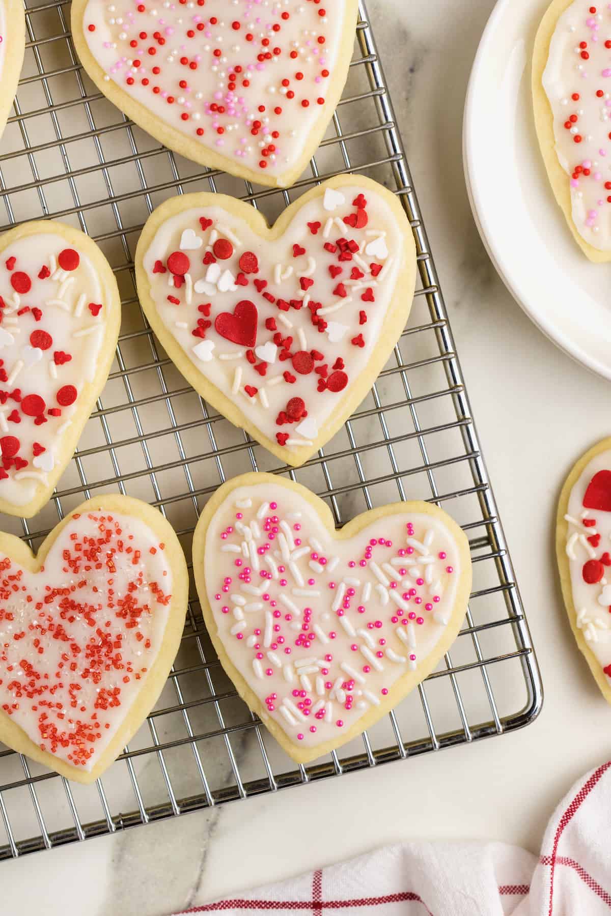 Heart shaped sugar cookies with white frosting and red and pink sprinkles on a wire cooling rack on a white marble counter.