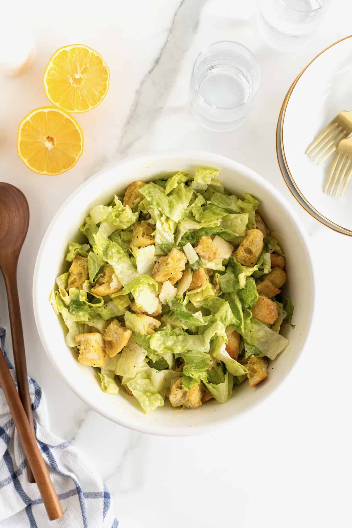A large white serving bowl filled with caesar salad topped with croutons. Two wooden spoons sit next to the bowl of salad.