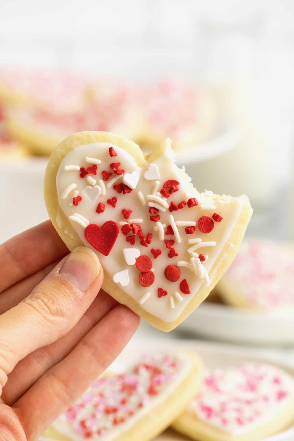 A hand holding a heart shaped sugar cookie with red and white sprinkles with a bite out of the top right corner.