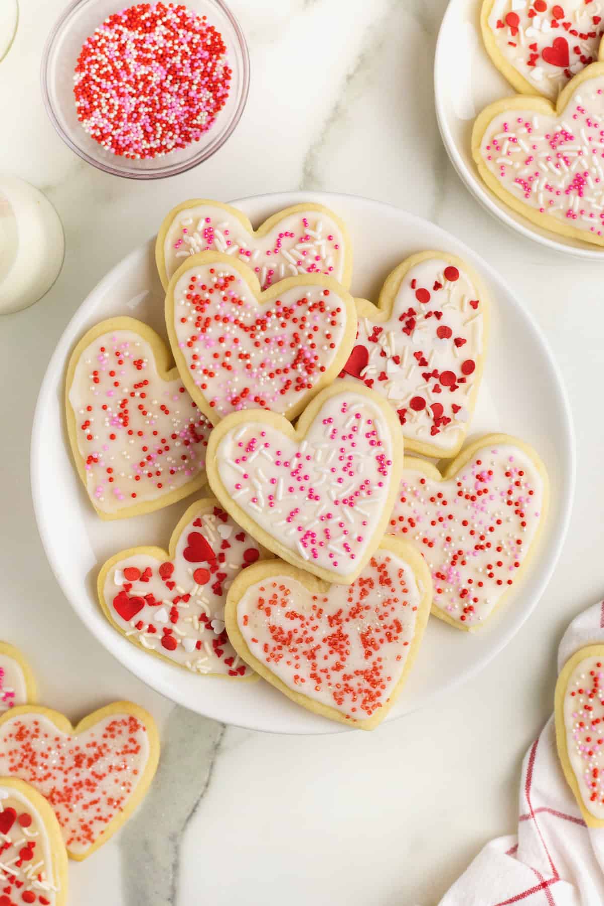 A pile of heart shaped sugar cookies with white frosting and pink and red sprinkles on a large white serving plate.