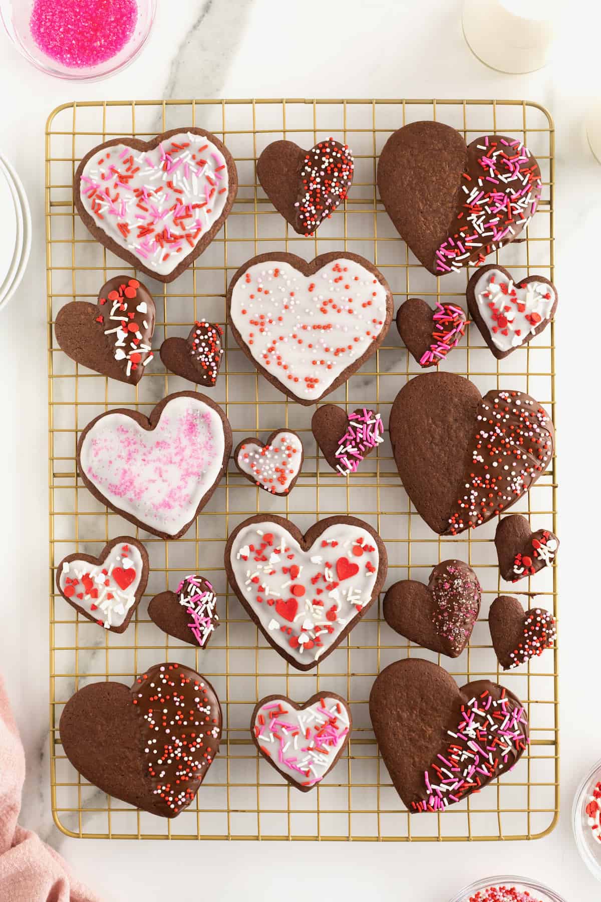A gold toned metal cooling rack with chocolate heart shaped cookies of various sizes. Some of the cookies have white frosting and pink and red sprinkles. Some have chocolate frosting with sprinkles.