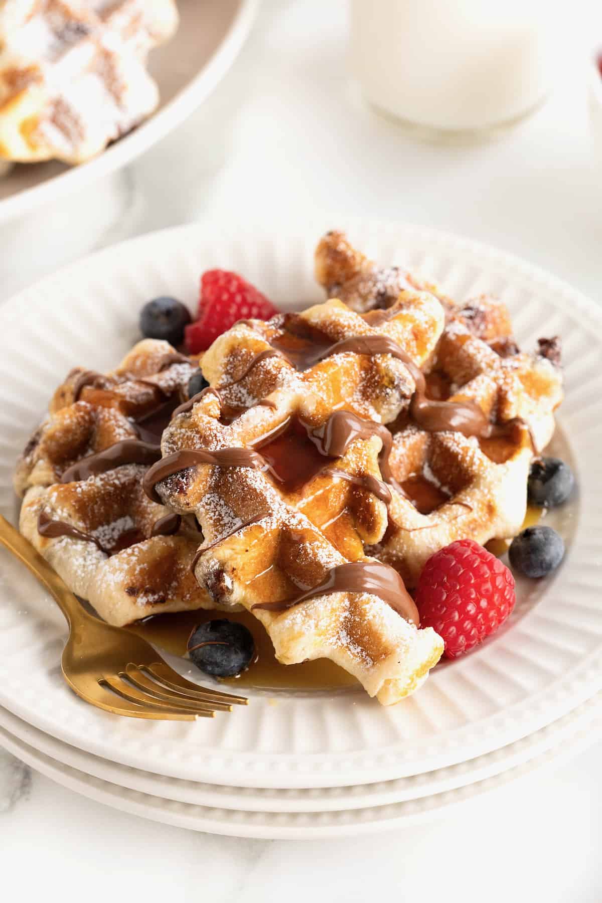 A large white fluted plate with two waffle-textured pastries topped with powdered sugar, maple syrup, a Nutella drizzle, blueberries and strawberries.